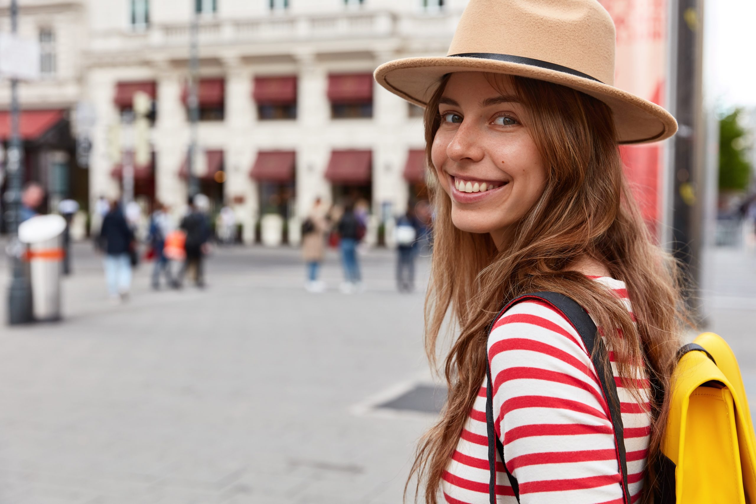 Horizontal view of delighted female tourist walks on street, wears stylish headgear and striped jumper, carries rucksack, poses over blurred urban setting with copy space for brand name or label