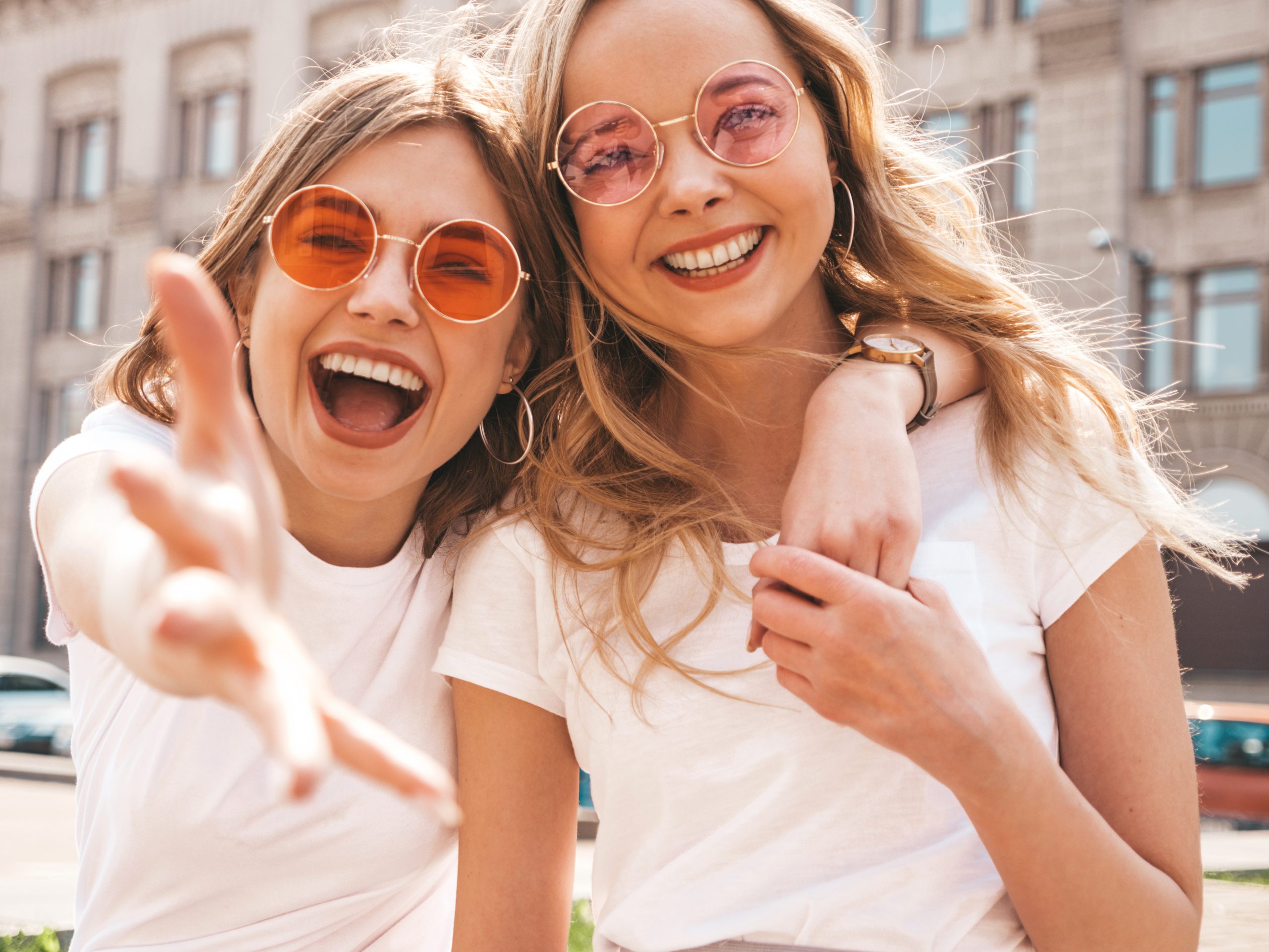 Portrait of two young beautiful blond smiling hipster girls in trendy summer white t-shirt clothes. Sexy carefree women posing on street background. Positive models having fun in sunglasses.Hugging